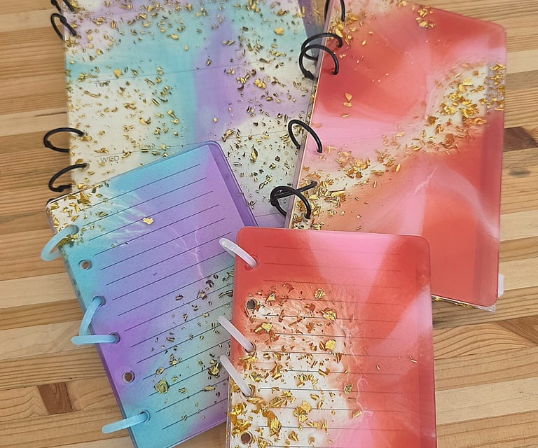 Carnets à anneaux aux couvertures en résine époxy pastel rehaussées de feuille d’or, posés sur un plan de travail en bois